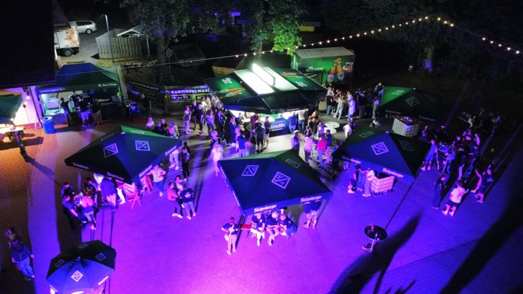 Das Foto zeigt das Spießbratenkonzert am Abend. Mehrere große Schirme stehen auf dem Marktplatz zwischen vielen Menschen, die in Gruppen beisammenstehen. In der Mitte befindet sich ein Bierbrunnen. Die Beleuchtung ist teils lila und teils blau.